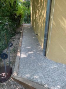 Allée en gravier clair longeant un mur jaune avec une bordure en bois dans un jardin ombragé.