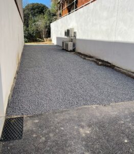Allée de jardin recouverte de gravier gris entre des murs blancs, avec climatiseurs extérieurs en arrière-plan.