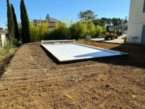 Piscine avec volet roulant blanc sur terrain en cours d'aménagement paysager avec pelleteuse et cyprès.