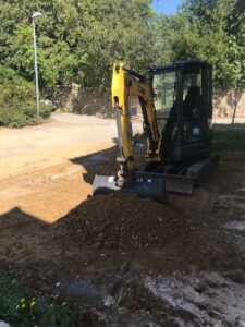 Mini-pelle jaune effectuant des travaux de terrassement sur un tas de terre dans un jardin ensoleillé.