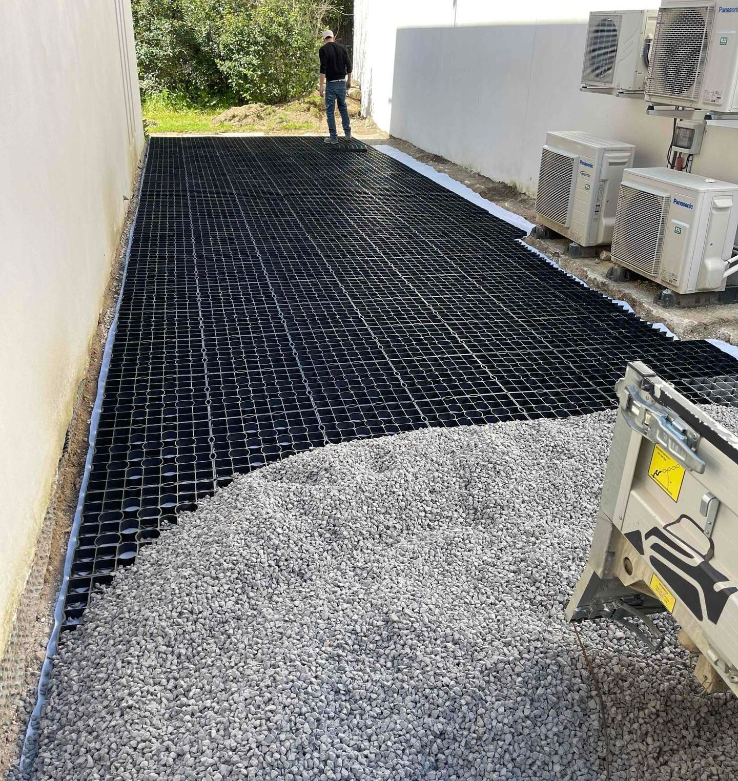 Gravier versé sur grilles de stabilisation noires dans une allée avec unités de climatisation Panasonic.