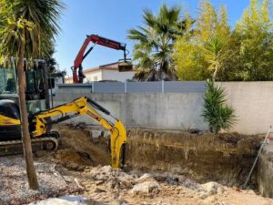 Mini-pelle jaune MIRAVET TP en pleine excavation et grue de déblaiement pour un terrassement de jardin avec palmiers.